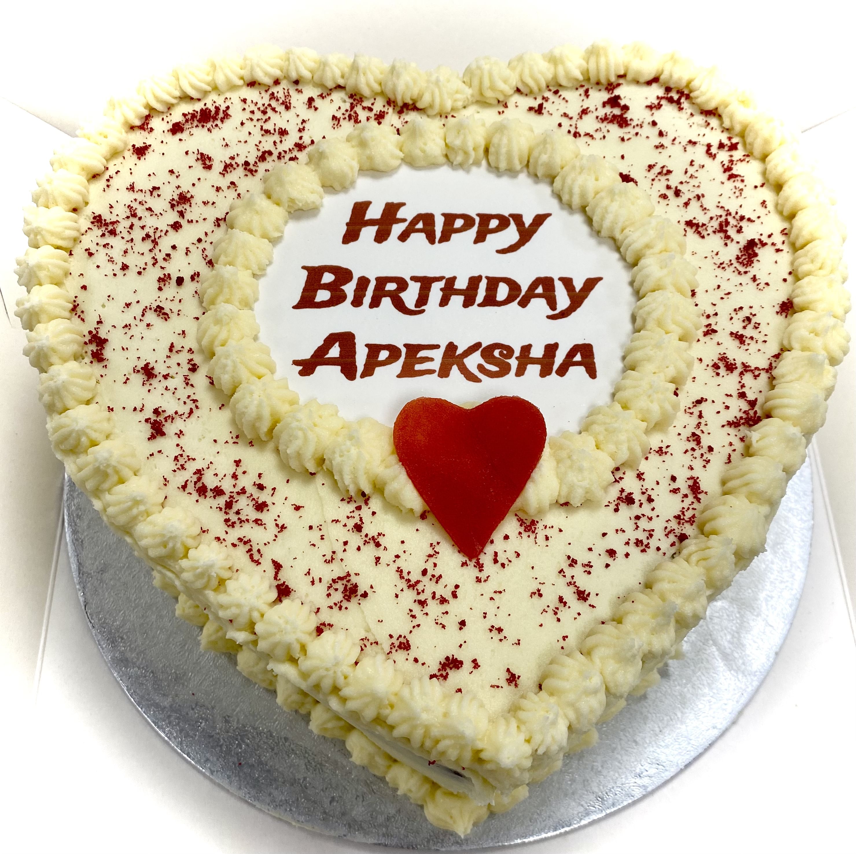 A heart-shaped cake with an elegant design features white frosting and red sprinkles. Adorning the top, a red heart decoration and the text 'Happy Birthday Apeksha' written in red make it perfect for any love celebration.